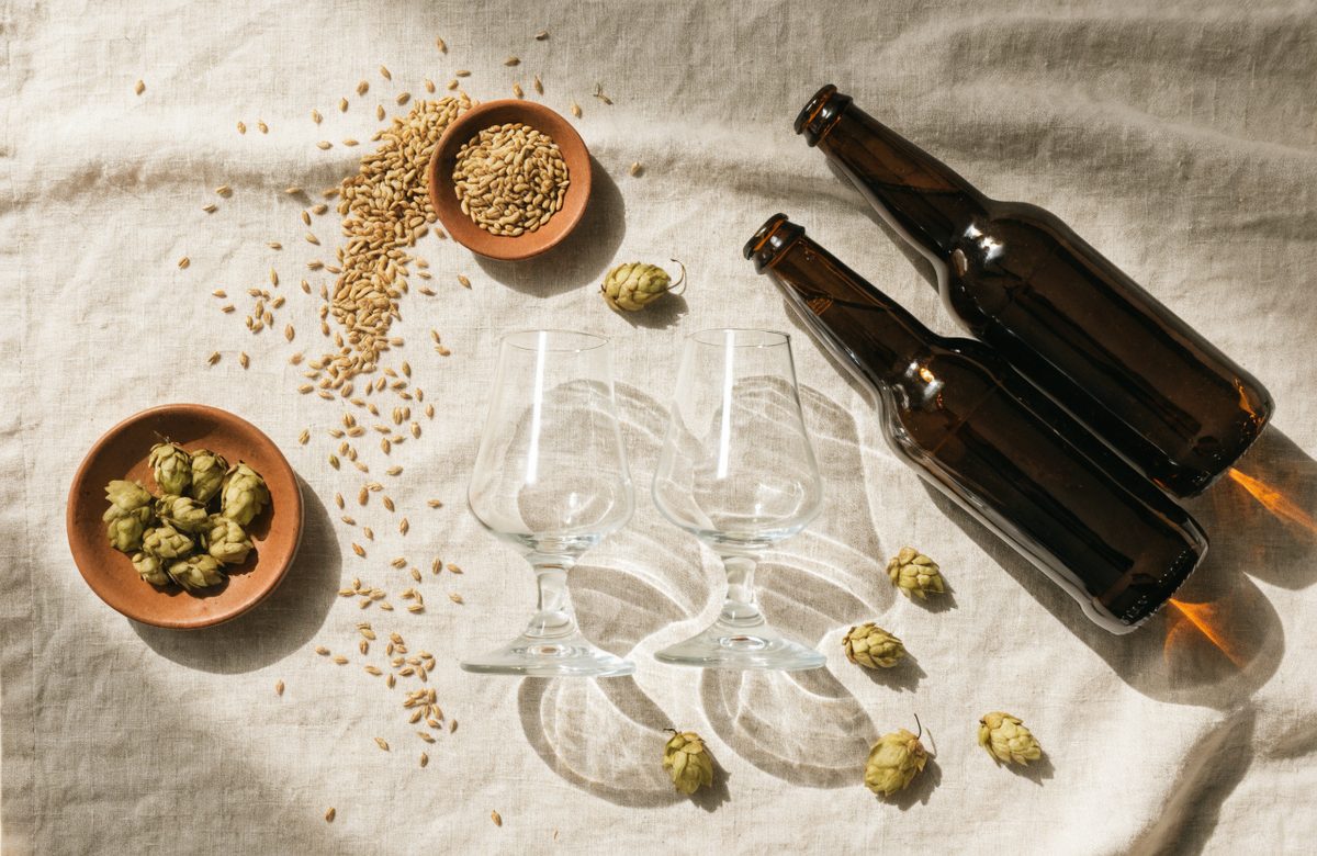 Flatlay mit zwei tulpenfoermigen Bierglaesern, dunklen Bierflaschen, Hopfendolden und Gerstenkoernern auf cremefarbenem Leinen in Creme- und Terracottatoenen