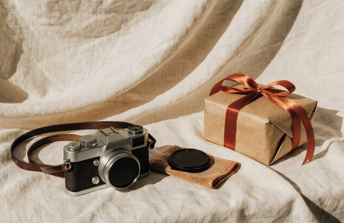 Stillleben mit Kamera, Lederkameragurt, Objektivdeckel, Mikrofasertuch und in Kraftpapier eingepacktem Geschenk mit Terracotta-Band auf cremefarbenem Leinen