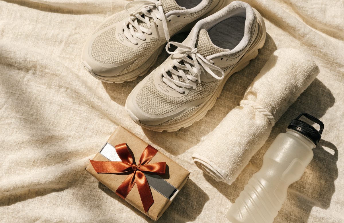 Flatlay mit Laufschuhen, gerolltem Handtuch, Trinkflasche und in Kraftpapier verpacktem Geschenk mit Terracotta-Schleife auf cremefarbenem Leinen