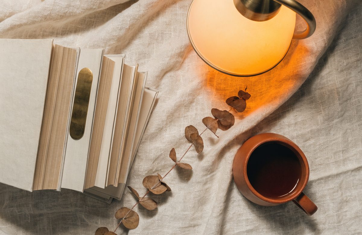 Flatlay mit einem Stapel Hardcover-Buecher, Messing-Lesezeichen, Leselampe und Terracotta-Tasse auf cremefarbenem Leinen