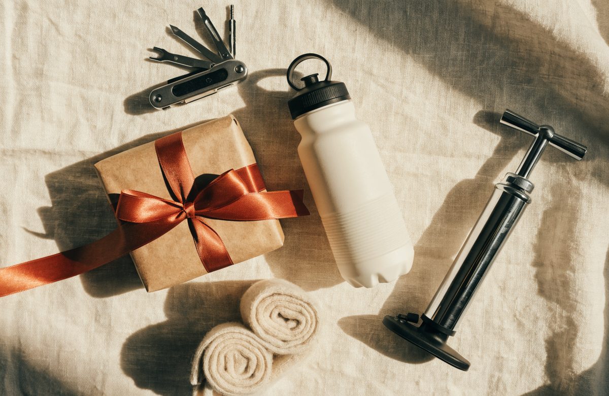 Flatlay mit Fahrrad-Multitool, isolierter Trinkflasche, Merinosocken, Minipumpe und in Kraftpapier verpacktem Geschenk mit Terracotta-Schleife auf cremefarbenem Leinen