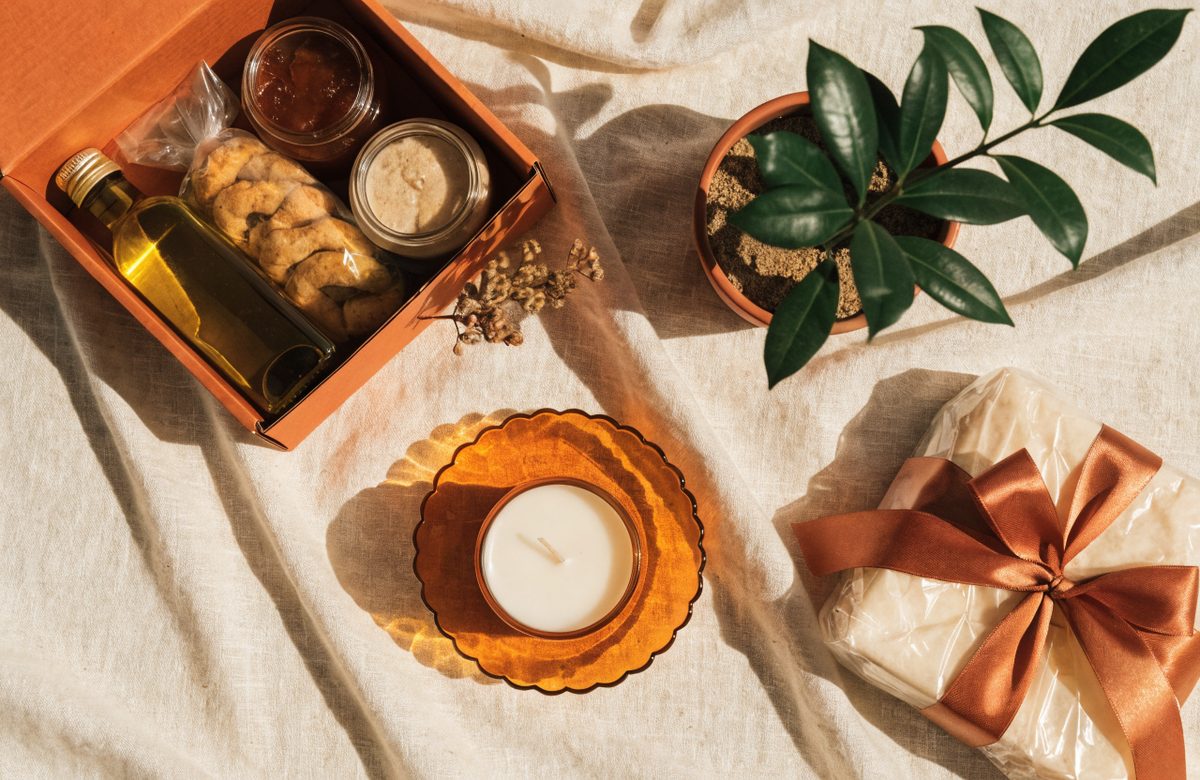 Elegant arrangiertes Geschenk mit Feinkost, Duftkerze und Zimmerpflanze auf cremefarbenem Leinen in Creme- und Terracottatoenen, ohne Menschen