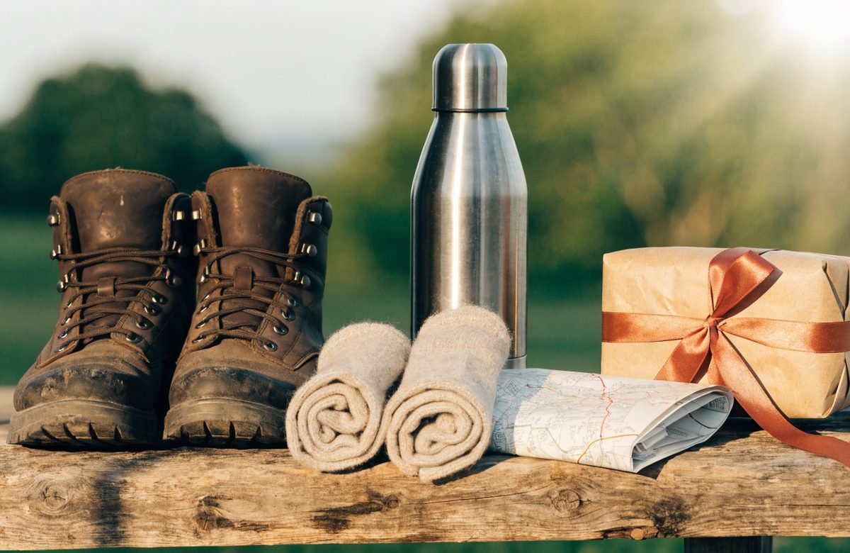 Stillleben mit Wanderstiefeln, aufgerollten Wollsocken, Thermosflasche, gefalteter Wanderkarte und in Kraftpapier eingepacktem Geschenk mit Terracotta-Band auf Holzbank