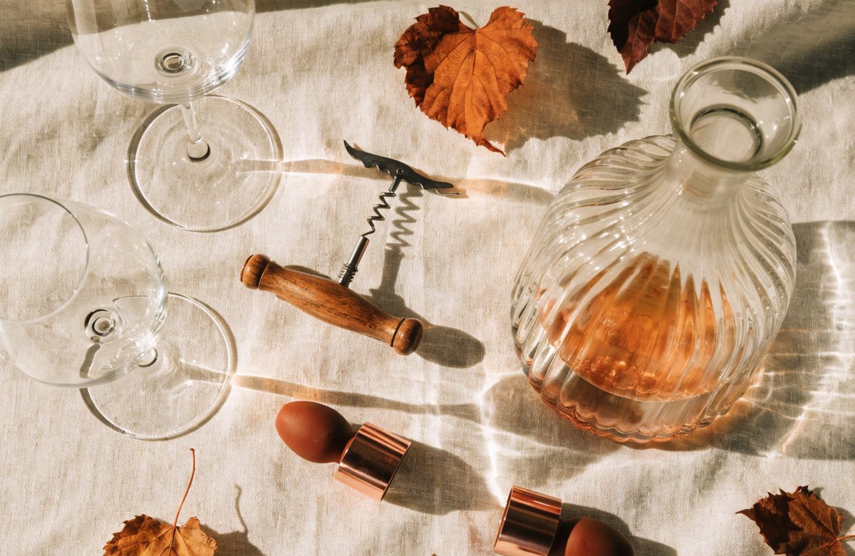 Geschenke für Weintrinker auf einem Holztisch: Dekantierkaraffe, zwei Gläser, Kellnermesser und Weinverschlüsse in warmen Creme- und Terrakotta-Tönen
