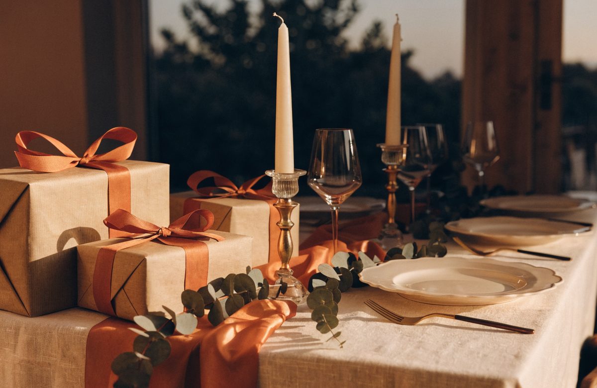 Festlich gedeckter Tisch mit in Kraftpapier verpackten Geschenken, Terracotta-Bändern, cremefarbenen Kerzen und Eukalyptuszweigen im warmen Abendlicht
