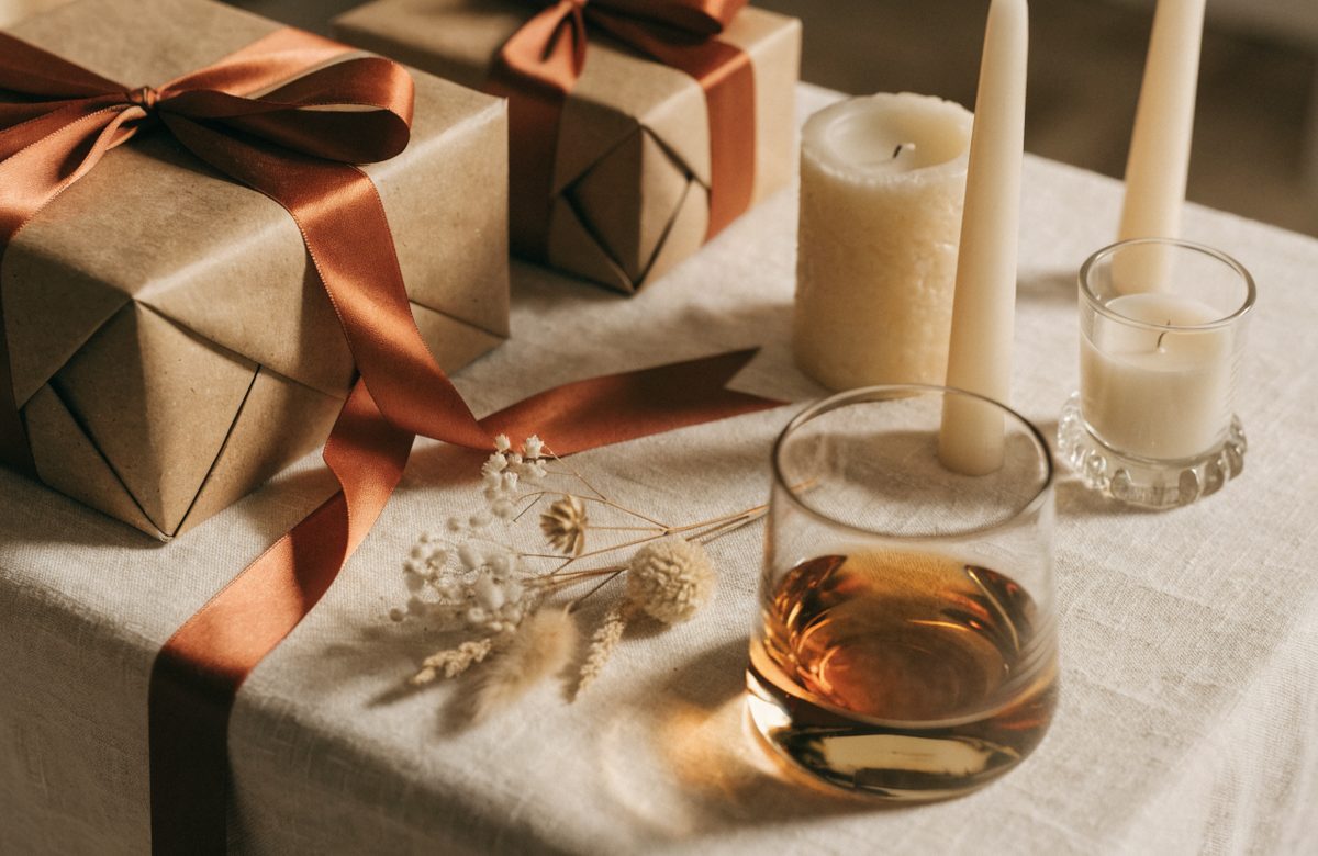 Festlich gedeckter Tisch mit in Kraftpapier verpackten Geschenken, Terracotta-Schleife, Kerzen, Whiskyglas und Trockenblumen auf cremefarbenem Leinen