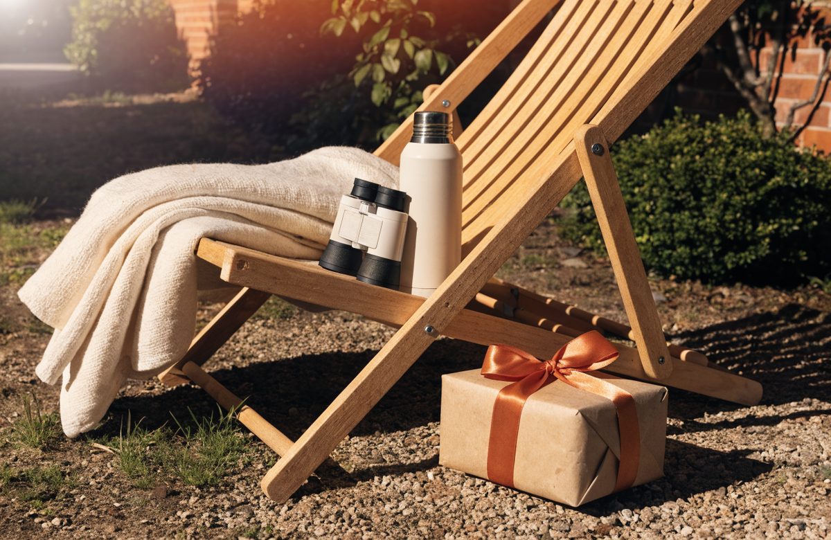 Stillleben mit Liegestuhl aus hellem Holz, gefalteter cremefarbener Decke, Fernglas, Thermoskanne und in Kraftpapier verpacktem Geschenk mit Terracotta-Schleife im warmen Gartenlicht
