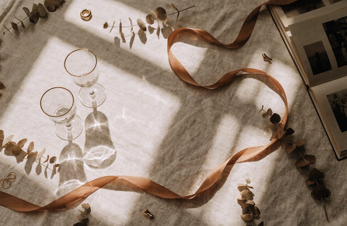 Flatlay zur goldenen Hochzeit auf cremefarbenem Leinen: zwei goldrandige Gläser, terrakottafarbenes Seidenband, Eukalyptuszweige, Eheringe und ein offenes Fotoalbum in warmem Licht