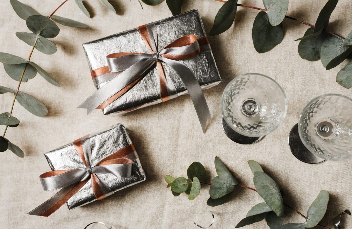Festlich verpackte Geschenke zur Silberhochzeit mit silbernem und terracottafarbenem Band, Sektgläsern und Eukalyptus auf cremefarbenem Leinen
